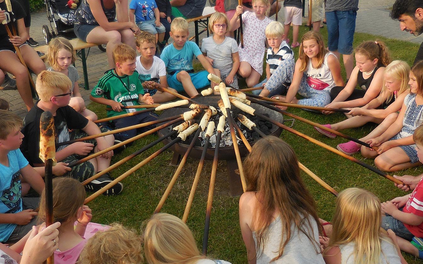 Kinder sitzen um Lagerfeuer uund halten Stockbrote ins Feuer
